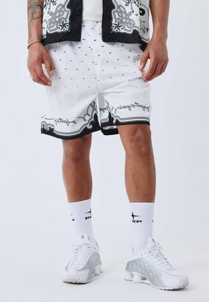 Witte shorts met zwarte accenten, voorzien van een decoratief patroon en tekst langs de zoom. Gekocht met witte sokken en lichtgrijze sneakers.