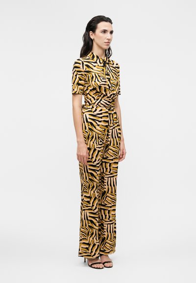 Γυναίκα στέκεται φορώντας ένα ταιριαστό πουκάμισο με κοντό μανίκι και παντελόνια με φαρδύ πόδι με καφέ, μαύρο και μπεζ αφηρημένο ζέβρα μοτίβο, μαύρες γόβες.