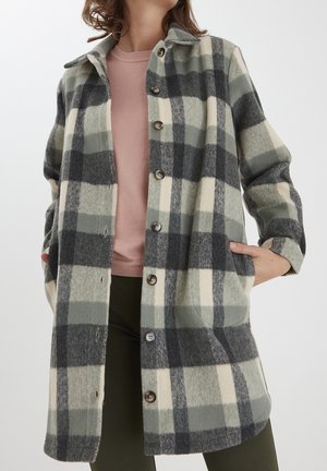Femme portant un long manteau en laine à carreaux noirs, gris et crème, par-dessus un haut rose et un pantalon foncé, les mains dans les poches du manteau.