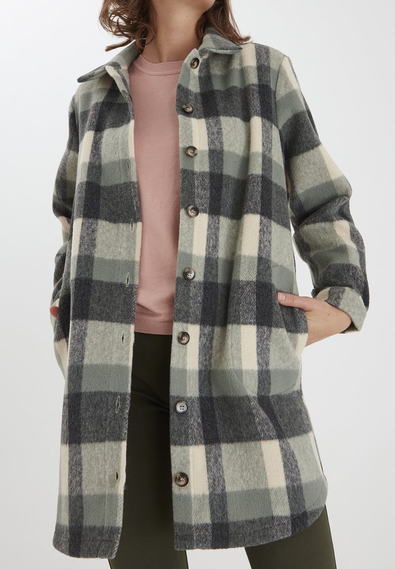 Femme portant un long manteau en laine à carreaux noirs, gris et crème, par-dessus un haut rose et un pantalon foncé, les mains dans les poches du manteau.