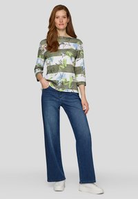 Gestreiftes Oberteil in mehreren Farben mit grünen, blauen und weißen Streifen sowie Sternmustern, kombiniert mit weit geschnittenen dunkelblauen Jeans und weißen Turnschuhen.