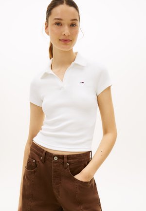 Mladá žena s hnedými vlasmi zviazanými do copu, oblečená v bielej polokošeli s krátkym rukávom a hnedých nohaviciach, s jednou rukou v vrecku.