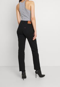 Jeans neri a vita alta con una vestibilità slim, dotati di tasche posteriori e un patch in pelle marrone. Abbinati a un top grigio senza maniche e tacchi neri.