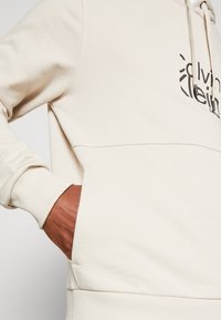 Lichtbeige hoodie met ribbels aan de mouwen en een voorzak. Beschikt over een zwart logo op de borst. Zachte stof met een gladde textuur.