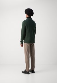 Jersey verde trenzado con cuello, combinado con pantalones beiges. La persona lleva zapatos negros y está de pie frente a un fondo liso.