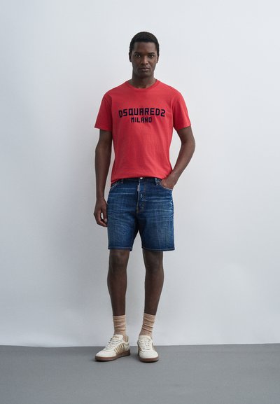 Άνδρας στέκεται μπροστά σε απλό τοίχο, φοράει κόκκινο t-shirt DSQUARED2 Milano, μπλε τζιν σορτς, μπεζ κάλτσες και λευκά αθλητικά παπούτσια με χρυσές ρίγες.