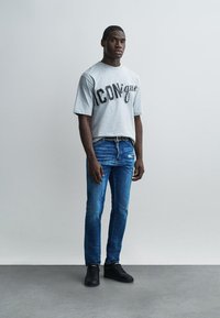 Άνδρας που στέκεται μπροστά σε απλή τοίχο φορώντας γκρι μπλουζάκι με το κείμενο "ICONique", μπλε τζιν, μαύρα αθλητικά παπούτσια και μαύρη ζώνη.