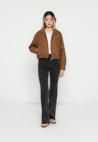 Veste bomber en daim marron avec fermeture éclair, poignets côtelés et coupe décontractée, associée à un jean noir évasé et des chaussures de couleur claire.