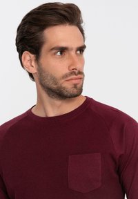 Burgundin pitkähihaisia paita, jossa on ribattu rakenne, pyöreä kaula-aukko ja vasemmassa rinnassa paikallinen tasku. Hihoissa on sileä pinta kontrastina.