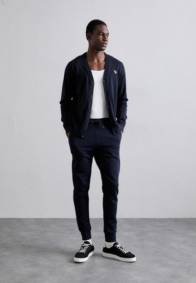 Άνδρας που στέκεται με τα χέρια στις τσέπες, φορώντας ναυτικό πλεκτό ζακέτα, λευκό μπλουζάκι, ναυτικές φόρμες, λευκές κάλτσες και μαύρα αθλητικά παπούτσια, μπροστά σε απλό τοίχο.