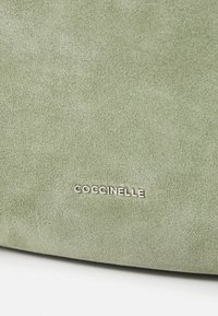 Zöld velúr kézitáska sima felülettel és finom fényességgel. Az anyagon kiemelkedő ezüst "Coccinelle" logó található.