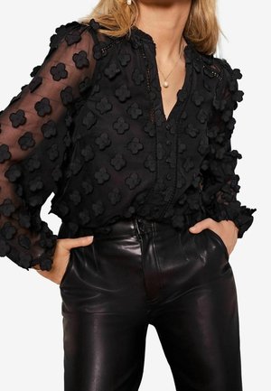 Vrouw die een doorschijnende zwarte blouse draagt met 3D bloemapplicaties en zwarte leren broek, handen in de zakken, met een parelketting en oorbellen.