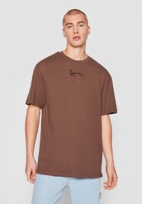 Homme aux cheveux courts portant un T-shirt marron uni oversize avec un texte brodé noir, associé à un jean bleu clair, sur fond gris.