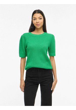 Mujer con cabello largo y oscuro, vestida con un suéter verde de manga corta y textura, y vaqueros negros de cintura alta, de pie sobre un fondo blanco.