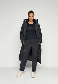 Black long puffer coat with a zippered front, paired with a fitted black top and loose black pants, worn with light beige sneakers.