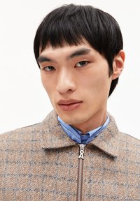 Jonge man met kort zwart haar, gekleed in een beige geruite zip-up jack en een blauwe blouse, tegen een effen achtergrond.