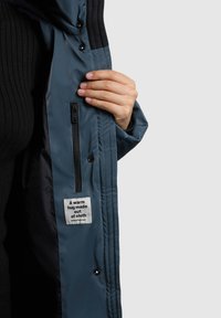 Modrá bunda s hladkým vnějším povrchem, černou fleecovou vnitřní částí, postranní kapsou na zip a látkovou štítkem s textem. Zapínání na zatrhávací knoflíky.