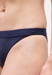 Κοντινό πλάνο της μέσης με σκουρόχρωμο μπλε κάτω μέρος μπικίνι με λεπτή μεταλλική λάμψη σε ανοιχτόχρωμο τόνο δέρματος.