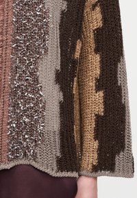 Κοντινό πλάνο σε μανίκι πουλόβερ πλεκτό στο χέρι με πολύχρωμα καφέ, μπεζ και γκρι νήματα που δημιουργούν υφή και μοτίβο.