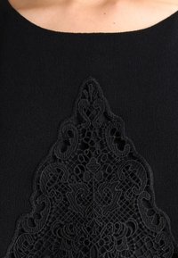 Prenda negra con intrincados bordados de encaje negro en un patrón decorativo, con un fondo de tela suave que contrasta con la textura del encaje.