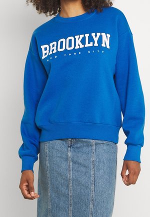 Personne portant un sweat-shirt bleu avec le texte "BROOKLYN NEW YORK CITY" et une jupe en denim bleu clair taille haute.