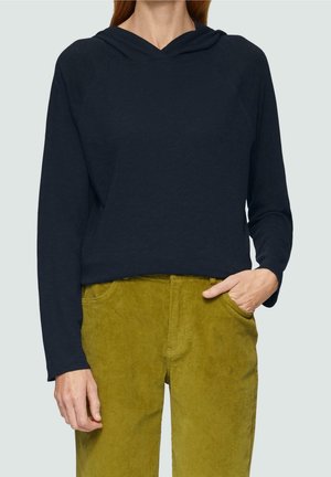 Persoon draagt een marineblauwe hoodie met lange mouwen, met één hand in de zak van mosterdgele corduroy broek, tegen een effen achtergrond.