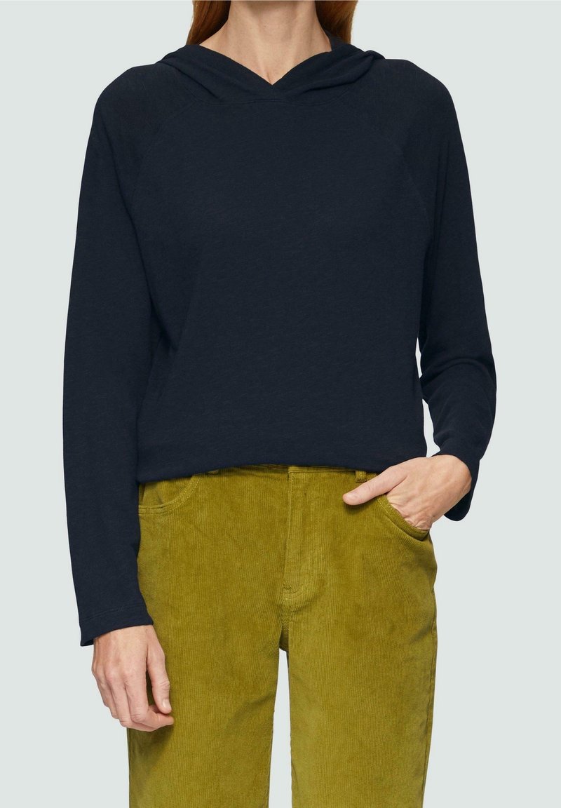 Osoba ubrana w granatową bluzę z kapturem z długim rękawem, z jedną ręką w kieszeni musztardowych sztruksowych spodni, na jednolitym tle.