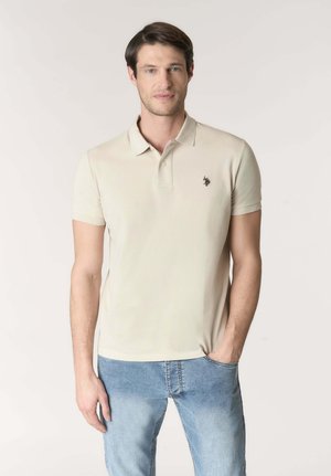 Uomo in piedi, indossa una polo beige a maniche corte con un piccolo logo nero ricamato e jeans azzurri.