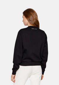 Sudadera negra con cuello redondo y mangas largas. Presenta "EST.1981" en relieve en blanco en la parte posterior. Textura suave, corte casual.