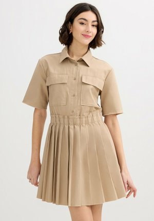 Mujer con vestido beige de manga corta con abertura de botones en la parte delantera, dos bolsillos en el pecho y falda plisada, posando frente a un fondo claro.