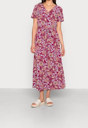 Robe maxi florale de couleur rose foncé avec des motifs paisley et des fleurs blanches. Encolure en V et manches courtes. Portée avec des sandales ouvertes de couleur claire.