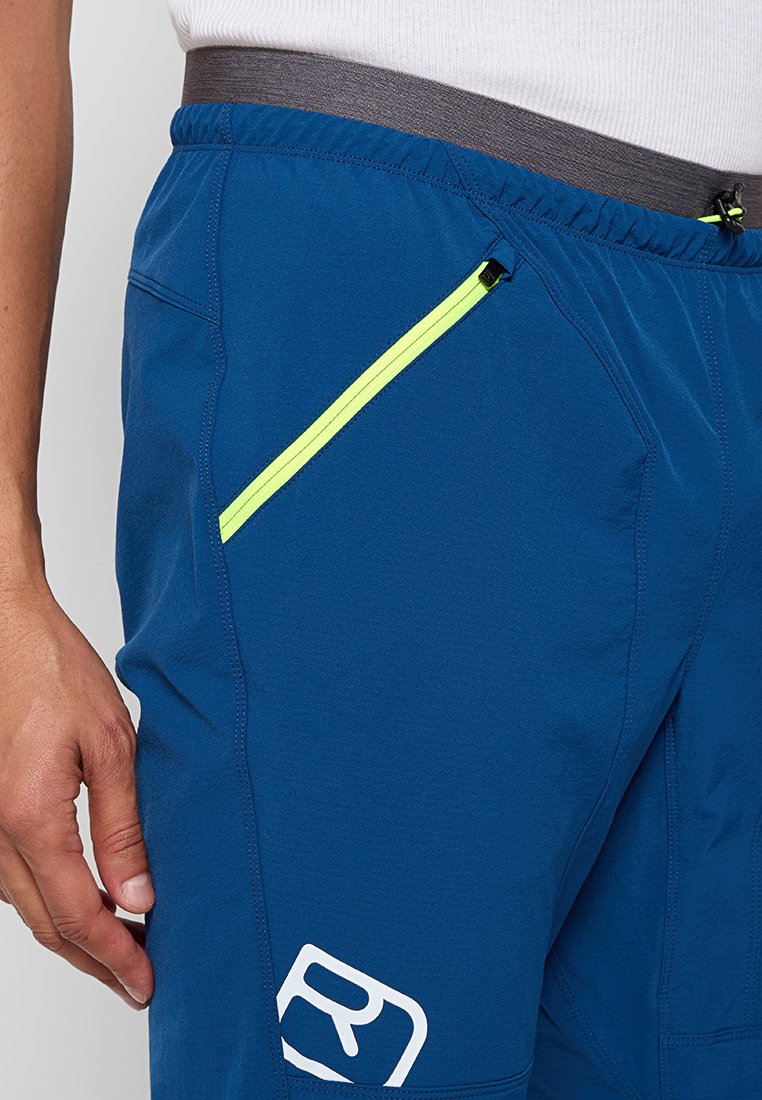 Gros plan sur une personne portant un short de sport bleu avec une poche zippée diagonale jaune fluo et un logo blanc près de la cuisse.