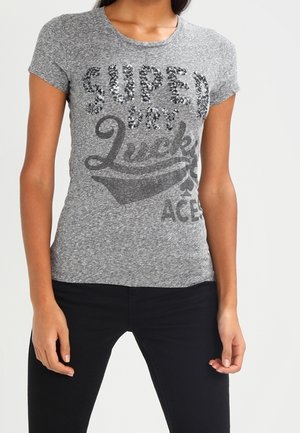 Kobieta w dopasowanej szarej koszulce z cekinowym napisem „SUPER DRY” oraz nadrukowanymi słowami „Lucky Aces”, połączona z czarnymi spodniami.