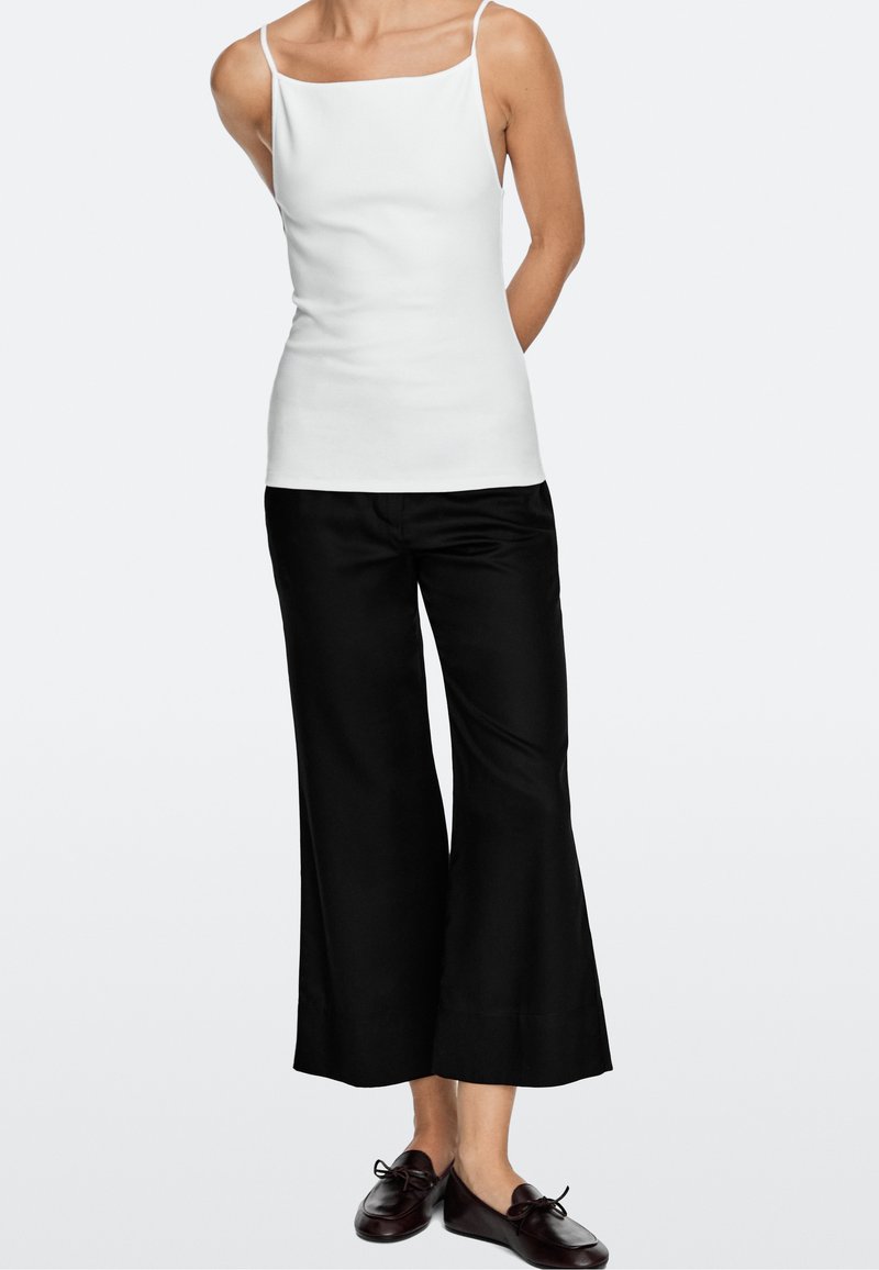 Persoon die een witte mouwloze top draagt, zwarte wijde cropped broek en donkerbruine leren loafers, staand met de handen achter de rug.