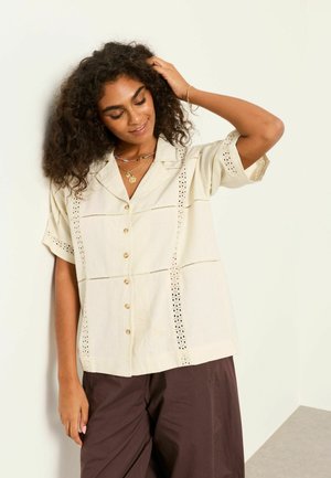Femme aux cheveux bouclés portant une chemise crème à manches courtes boutonnée avec des détails en dentelle et un pantalon marron foncé à jambes larges, touchant ses cheveux.
