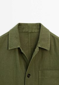 Chemise en lin vert olive avec col, coutures apparentes, bouton devant et poche poitrine sur fond blanc.