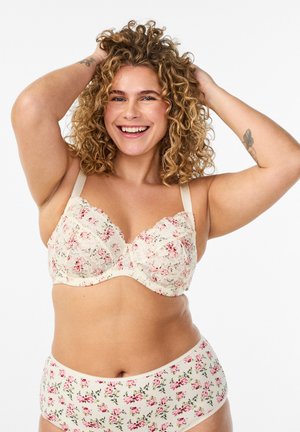 Žena s kučeravými vlasmi sa usmieva, ruky má vo vlasoch, oblečená v krémovej kvetinovej čipkovanej podprsenke a zodpovedajúcich vysokých nohavičkách na bielom pozadí.