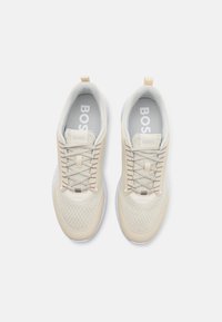 Lys beige idrettssko med mesh-overdel, rund tå og strukturert design. Har flate lisser og myk fòring med logo på sålen.