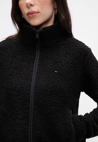 Svart fleecejakke med høy krage, glidelås og kontrasterende kant. Har en teksturert overflate og en liten logo.