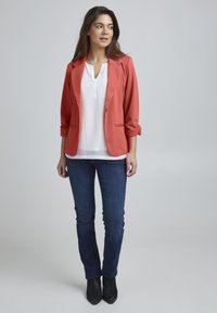Blazer corail avec les manches retroussées, poches avant et une chemise blanche contrastante en dessous ; associé à un jean bleu foncé et des bottines noires.