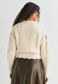 Cremefarvet kabelstrik sweater med bølget kant og teksturerede ærmer, parret med en mørkegrøn lædernederdel. Langt, krøllet hår er synligt.
