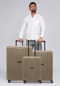 Drie lichtgewicht, beige harde koffers met een gestructureerde afwerking, voorzien van versterkte hoeken en uitschuifbare handvatten op wielen.