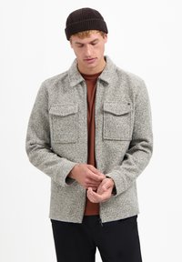 Grijze gestructureerde wollen jas met een kraag, twee borstzakken en een ritssluiting aan de voorkant. Draag het over een bruine shirt, gecombineerd met zwarte broek.