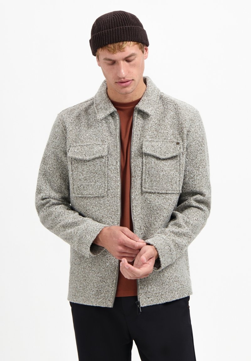 Grijze gestructureerde wollen jas met een kraag, twee borstzakken en een ritssluiting aan de voorkant. Draag het over een bruine shirt, gecombineerd met zwarte broek.