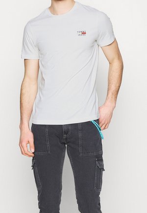 Mężczyzna ubrany w białą koszulkę z krótkim rękawem z logo "Tommy Jeans" oraz ciemnoszare spodnie cargo z niebieskim sznurkiem.