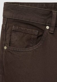 Gros plan sur la poche d’un jean en denim marron foncé avec coutures, rivets et passant de ceinture sur un fond clair.
