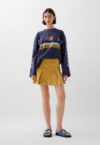 Lila langärmeliger Sweatshirt mit Grafik und Streifen, kombiniert mit einem gelben plissierten Jeansrock und blauen Sandalen mit metallen Akzenten.