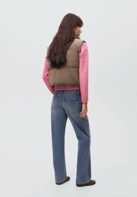 Gilet imbottito verde oliva su un maglione di maglia rosa, abbinato a jeans di denim azzurro chiaro. I capelli lunghi cadono lungo la schiena.