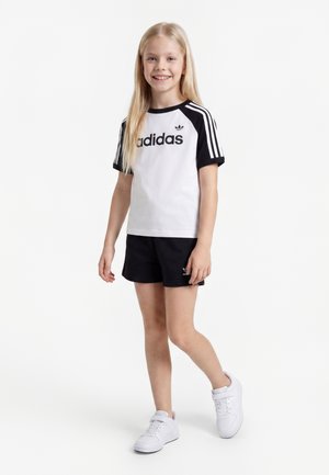 Jeune fille debout, portant un t-shirt Adidas blanc et noir, un short noir et des baskets blanches, souriante avec de longs cheveux blonds.