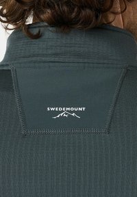 Mørkegrøn, tekstureret skjorte med krave. Hvidt "SWEDEMOUNT" logo med bjerggrafik på bagsiden. Synlige syede detaljer.